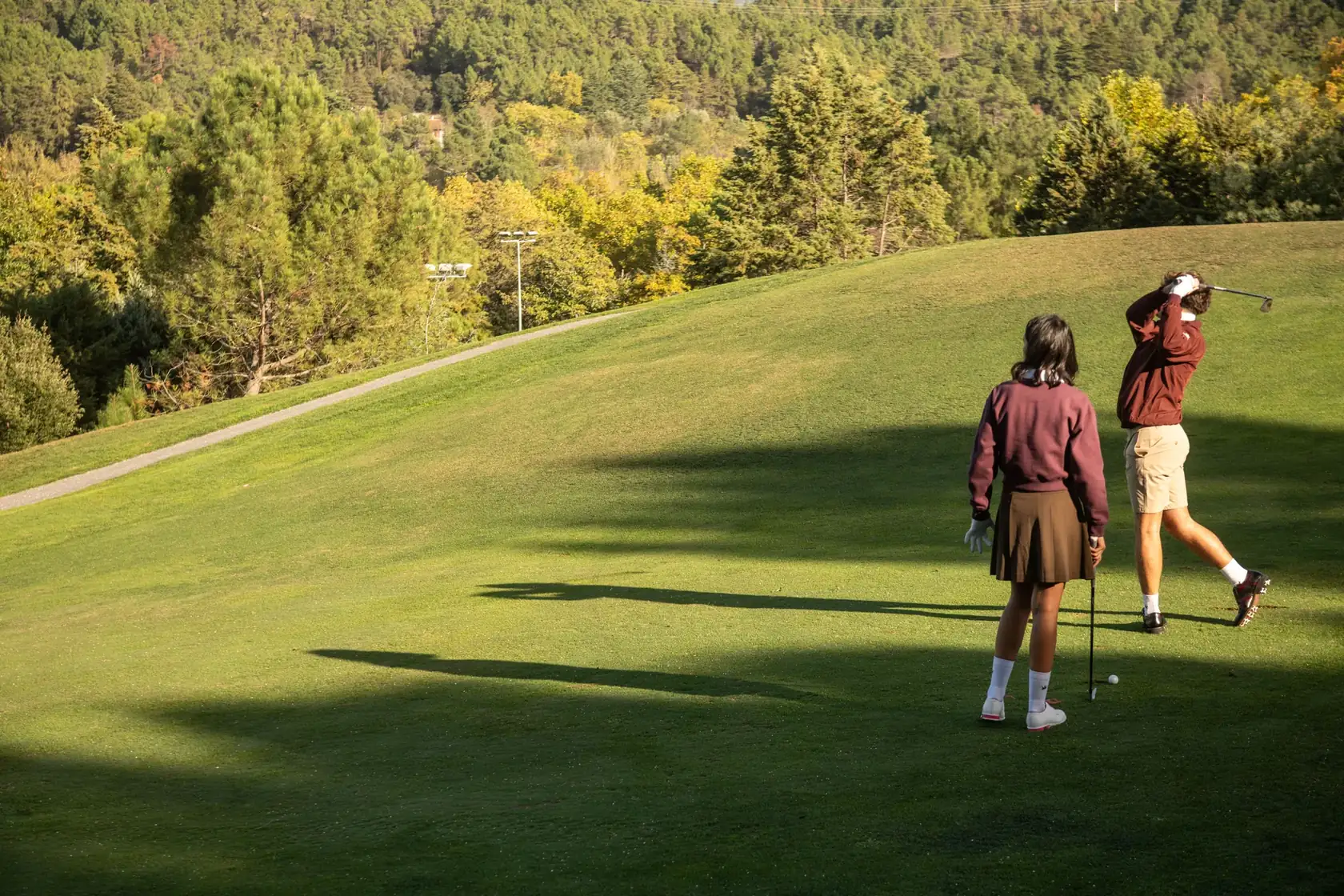 A golfer finishing his swing on a sunlit fairway in Lambda leather golf shoes, his partner watching