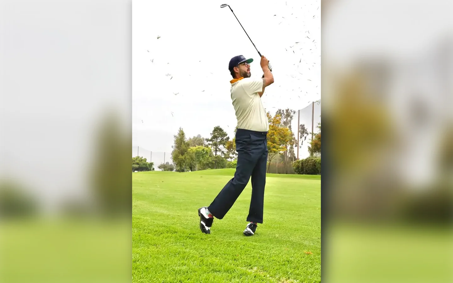 Golfer from Vintage Golf Wear in a cream polo, navy cap and high-waisted navy trousers finishing his swing on the fairway in black and white Lambda Golf shoes