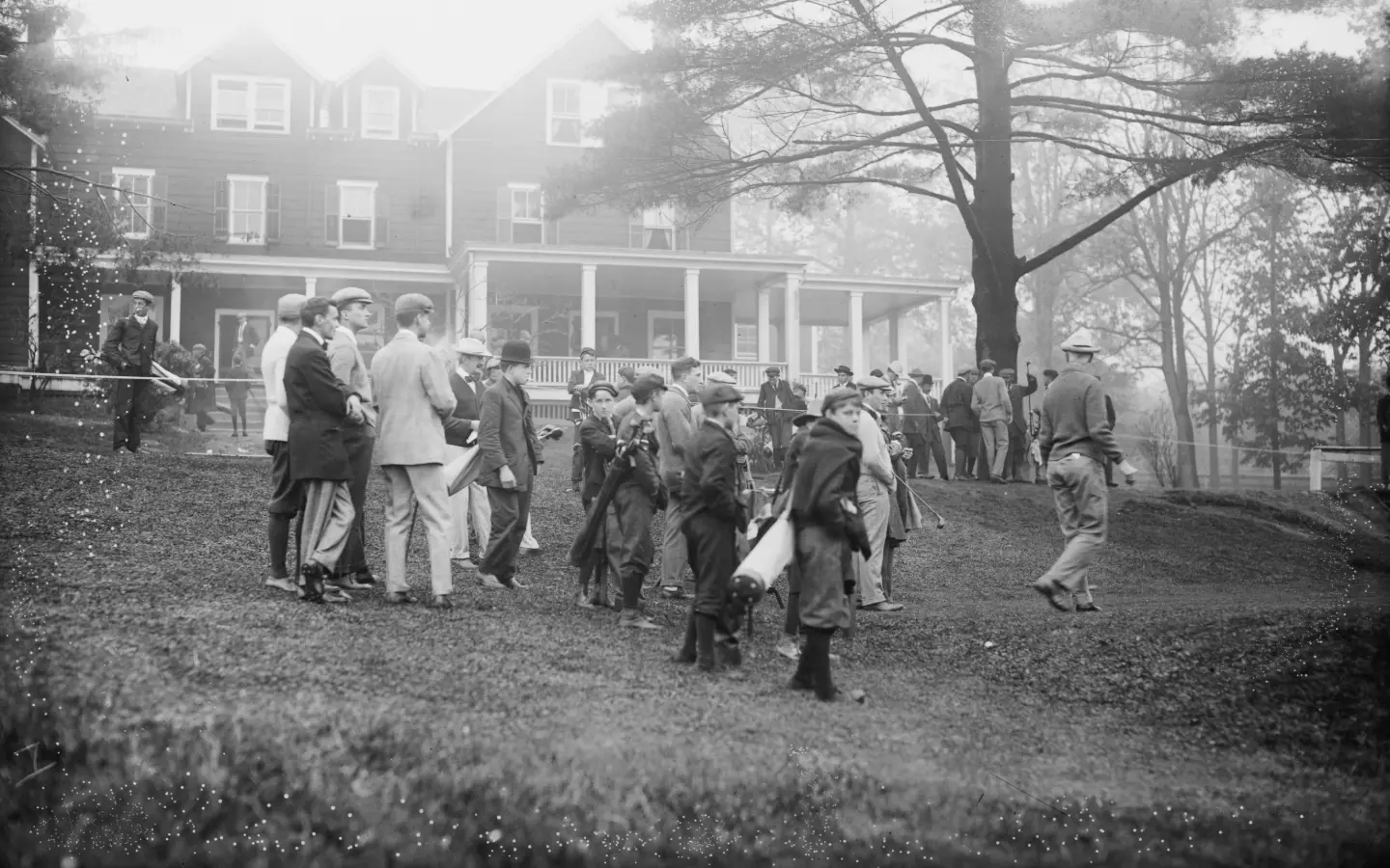 Black-and-white 1908 photograph of a crowd of spectators and golfers in hats and long coats gathered on the grass beside a clubhouse at a golf match at Baltusrol